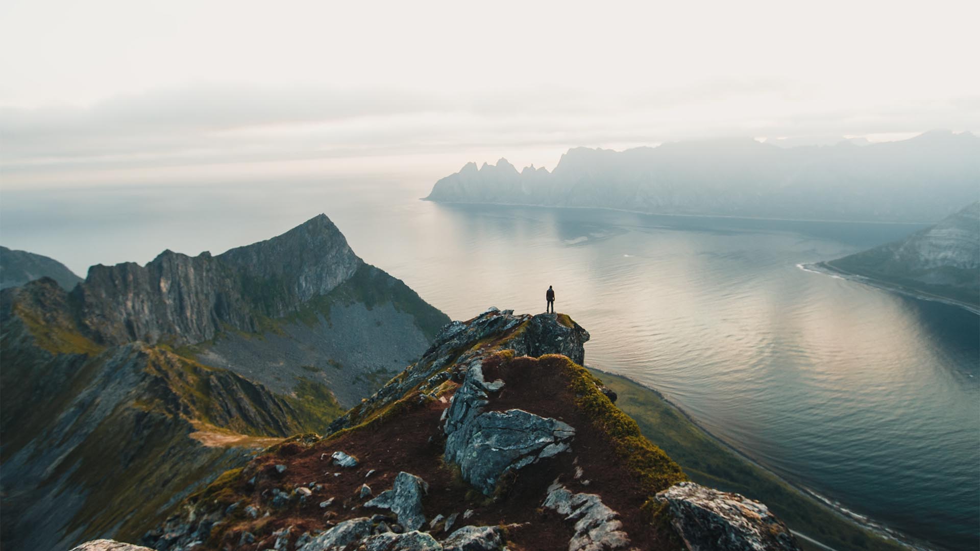 Mann auf Bergspitze mit Meer und Bergen im Hintergrund.